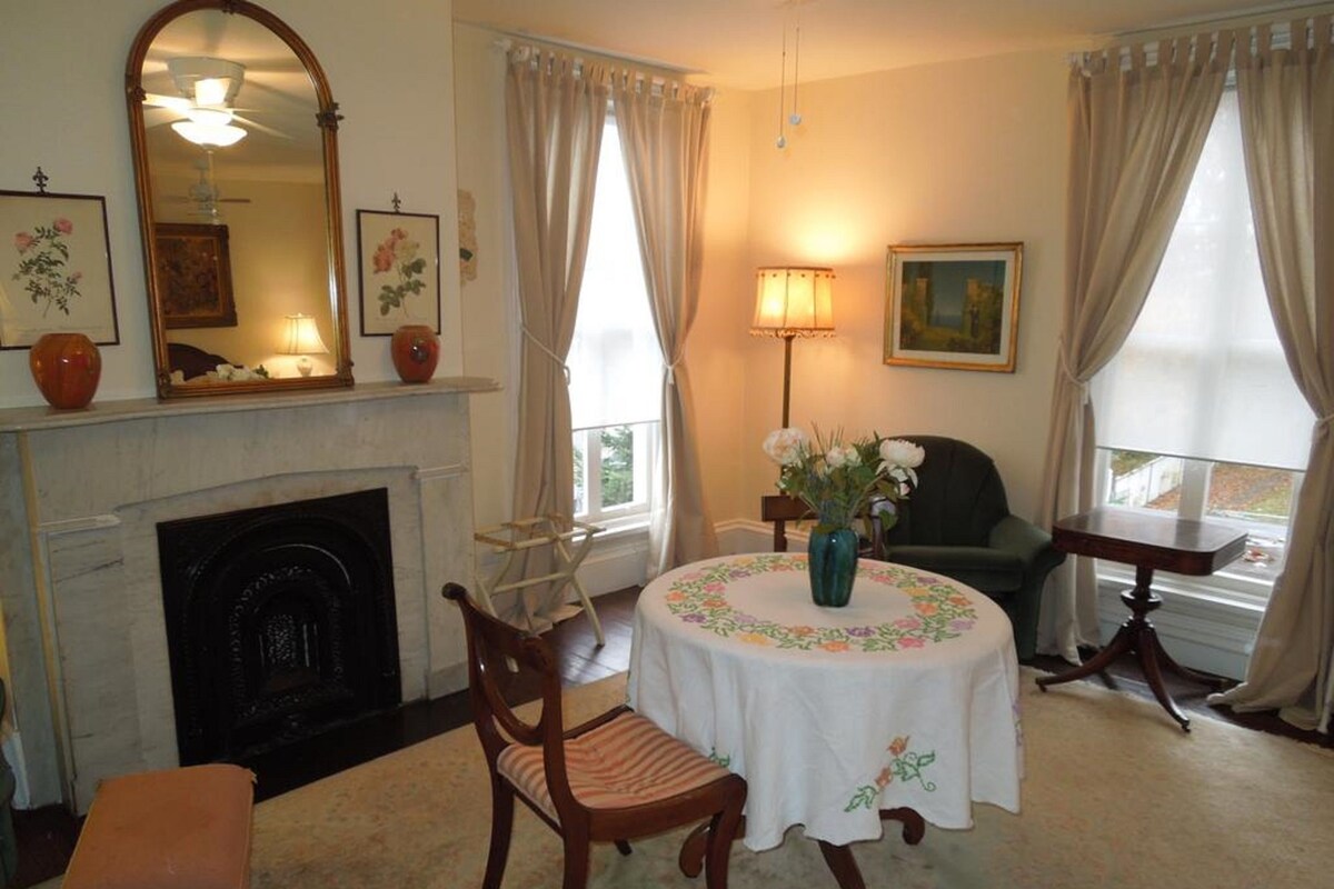 A room with a fireplace, a round table with a floral tablecloth, a chair, a small table with a lamp, and a painting on the wall.