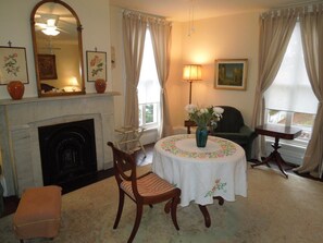 A room with a fireplace, a round table with a floral tablecloth, a chair, a small table with a lamp, and a painting on the wall. - Victorian Bed & Breakfast (Staten Island)