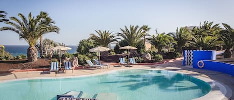 A swimming pool with lounge chairs and umbrellas, surrounded by palm trees and a view of the ocean.