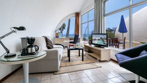 Un salon moderne doté d’un plafond incurvé, d’un canapé, d’une machine à café, d’un téléviseur et offrant une vue sur la mer grâce à de grandes fenêtres.
