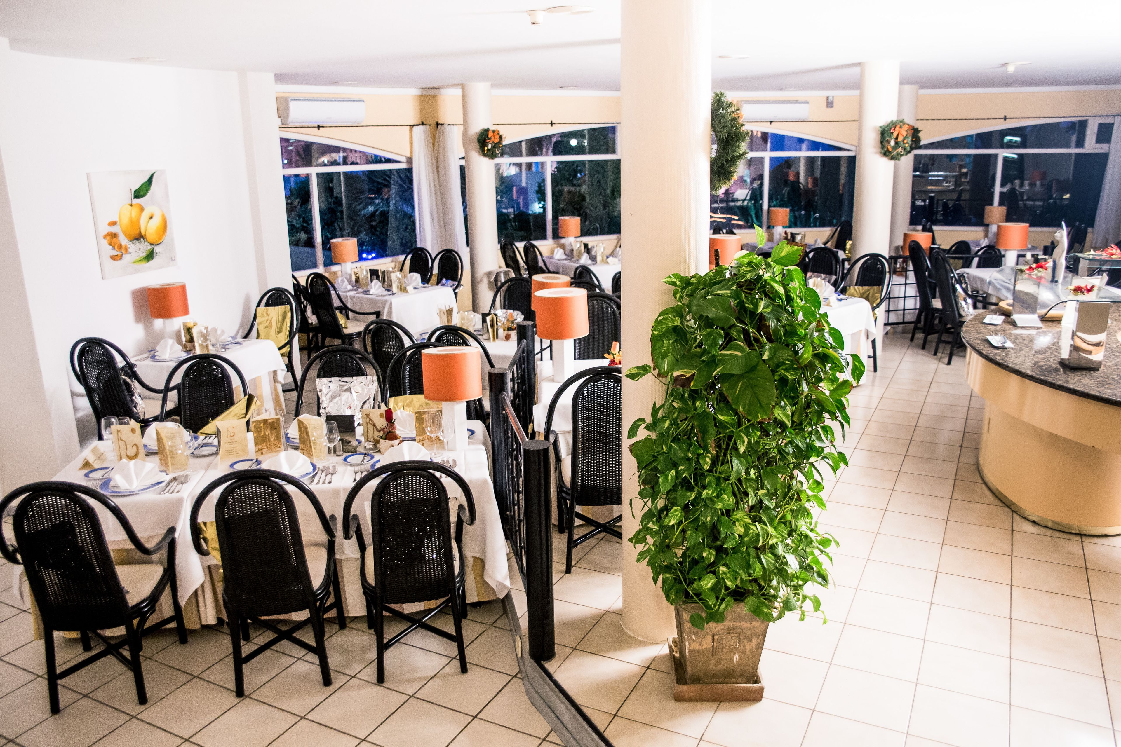 A restaurant with multiple tables, black chairs, and a large potted plant.