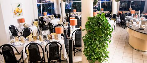 A restaurant with multiple tables, black chairs, and a large potted plant.