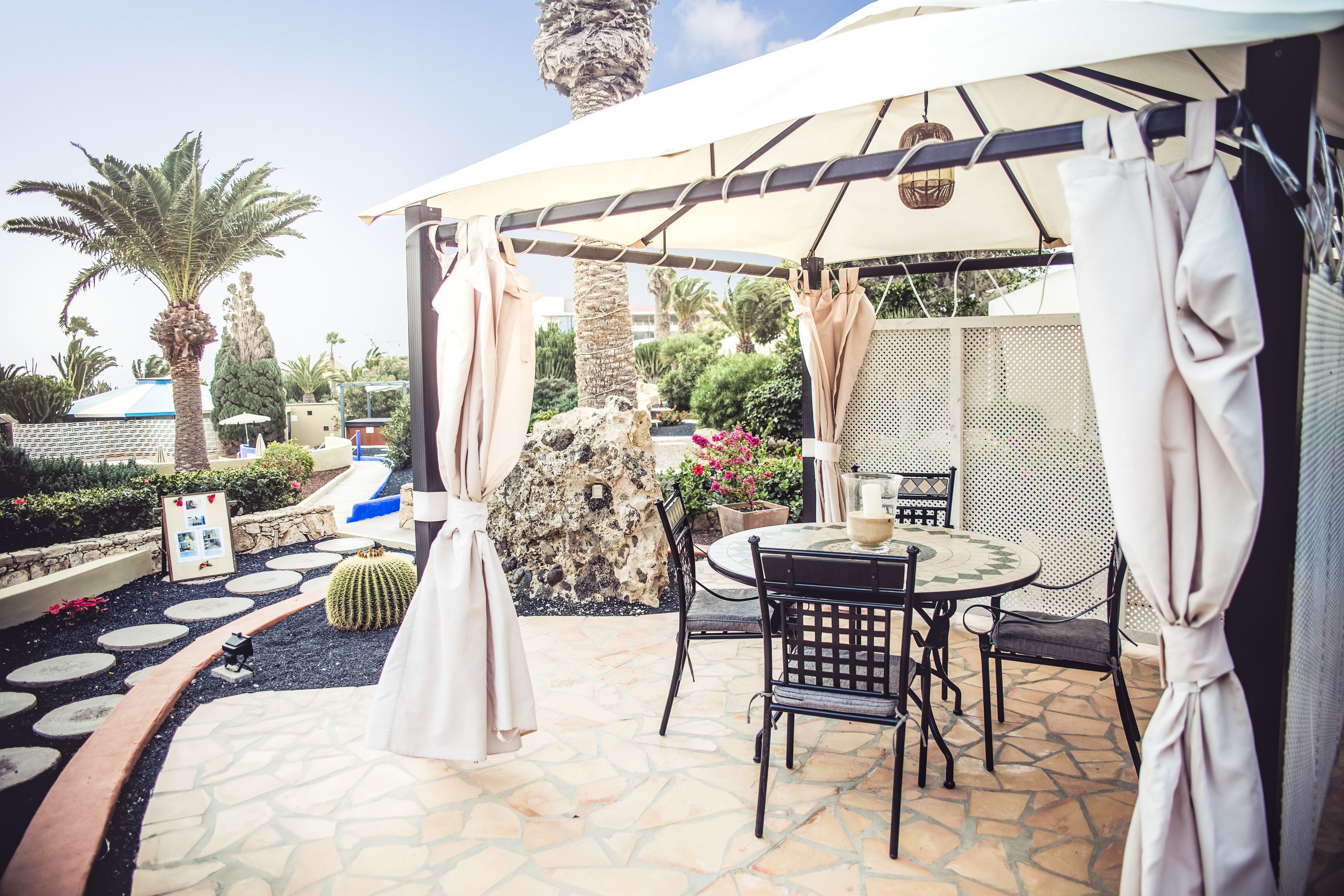 An outdoor patio area with a round table and chairs, a pergola with curtains, and a stone pathway.