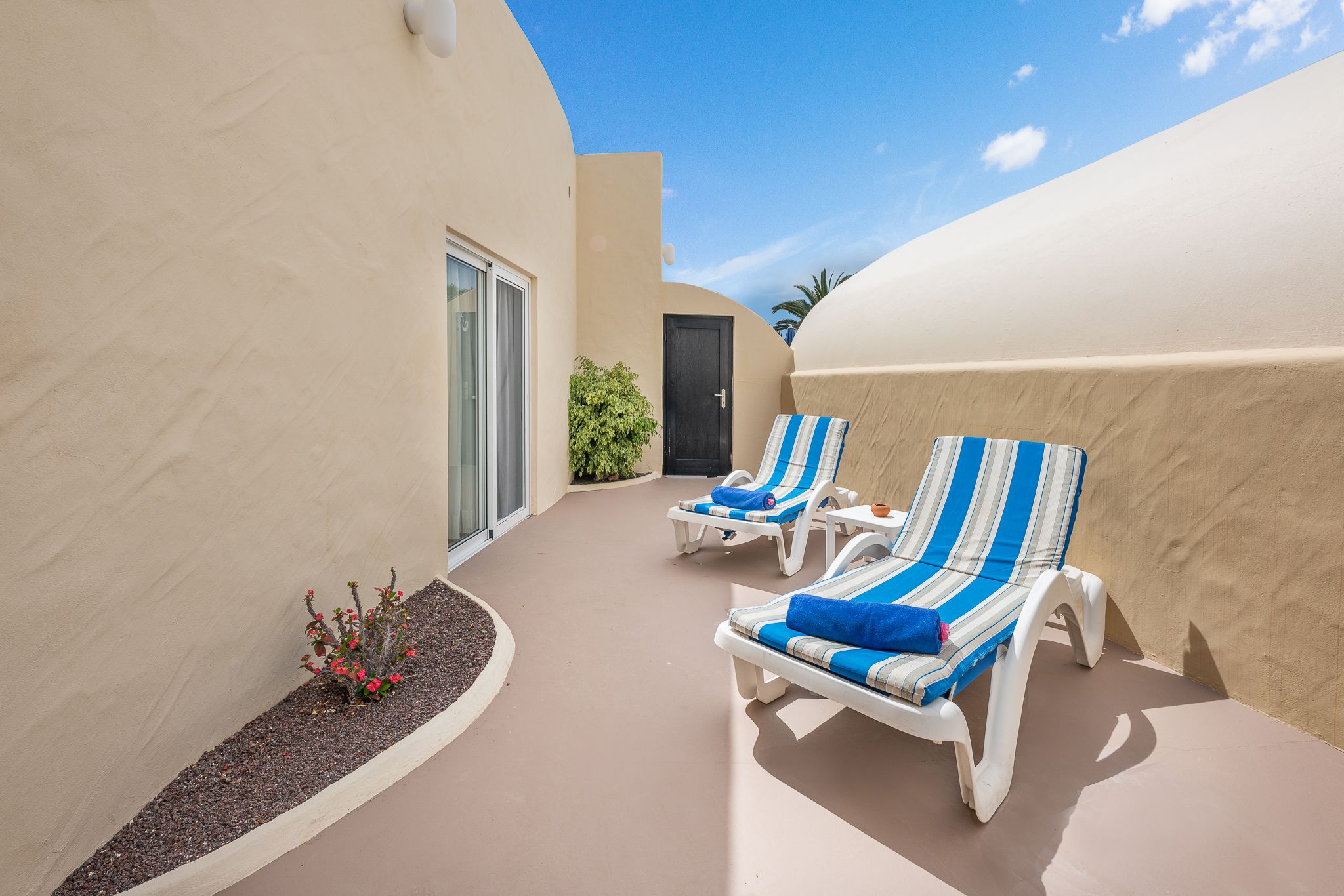 Een zonnig terras met twee witte loungestoelen, waarvan er een met een blauwe handdoek is uitgerust.