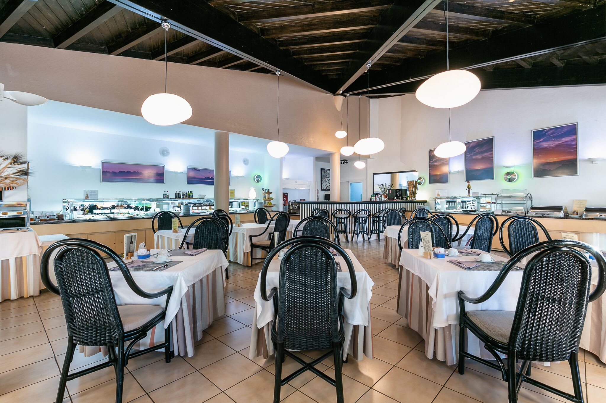 Un restaurant avec des tables rondes et des chaises en osier noir, des nappes blanches et des luminaires suspendus.