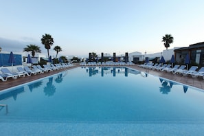 Una piscina con sillas de descanso y sombrillas, rodeada de palmeras y edificios.
