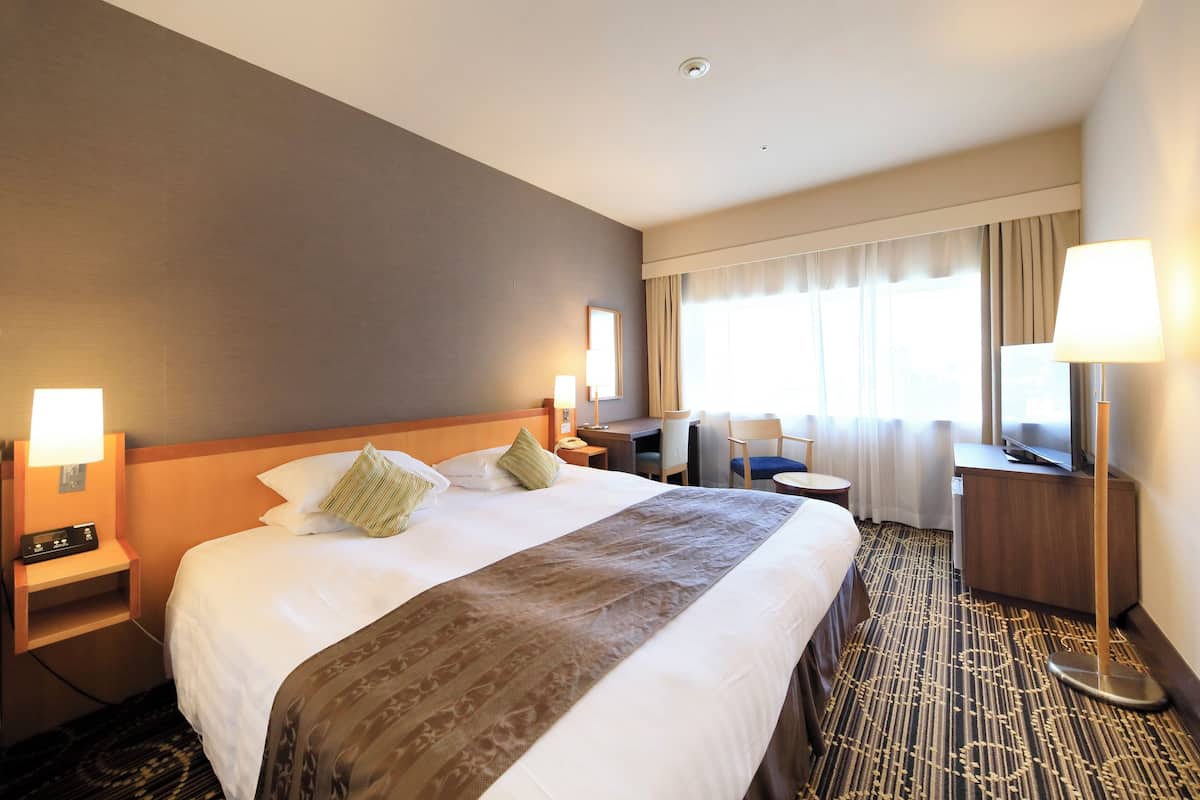 Quarto de hotel com uma cama grande, uma escrivaninha, uma cadeira, uma televisão e um abajur.