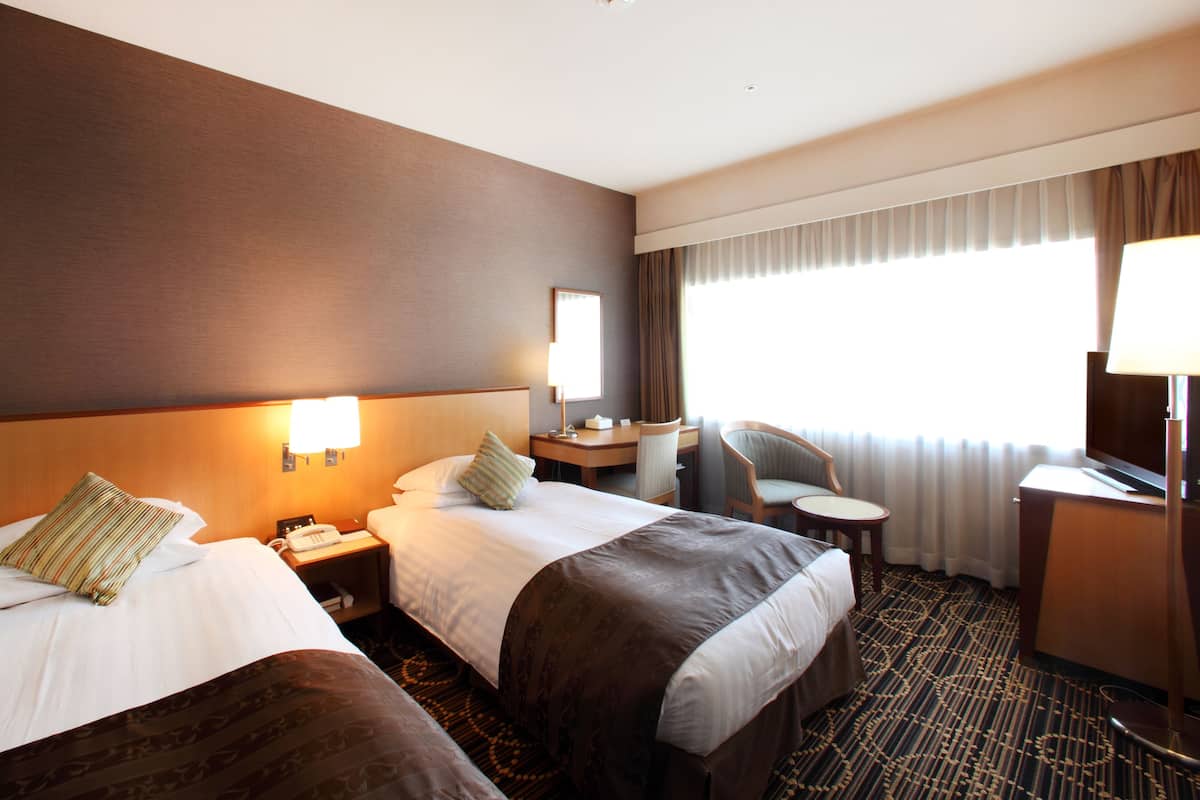 Quarto de hotel com duas camas, uma mesa, uma cadeira, uma luminária e uma janela grande com cortinas.