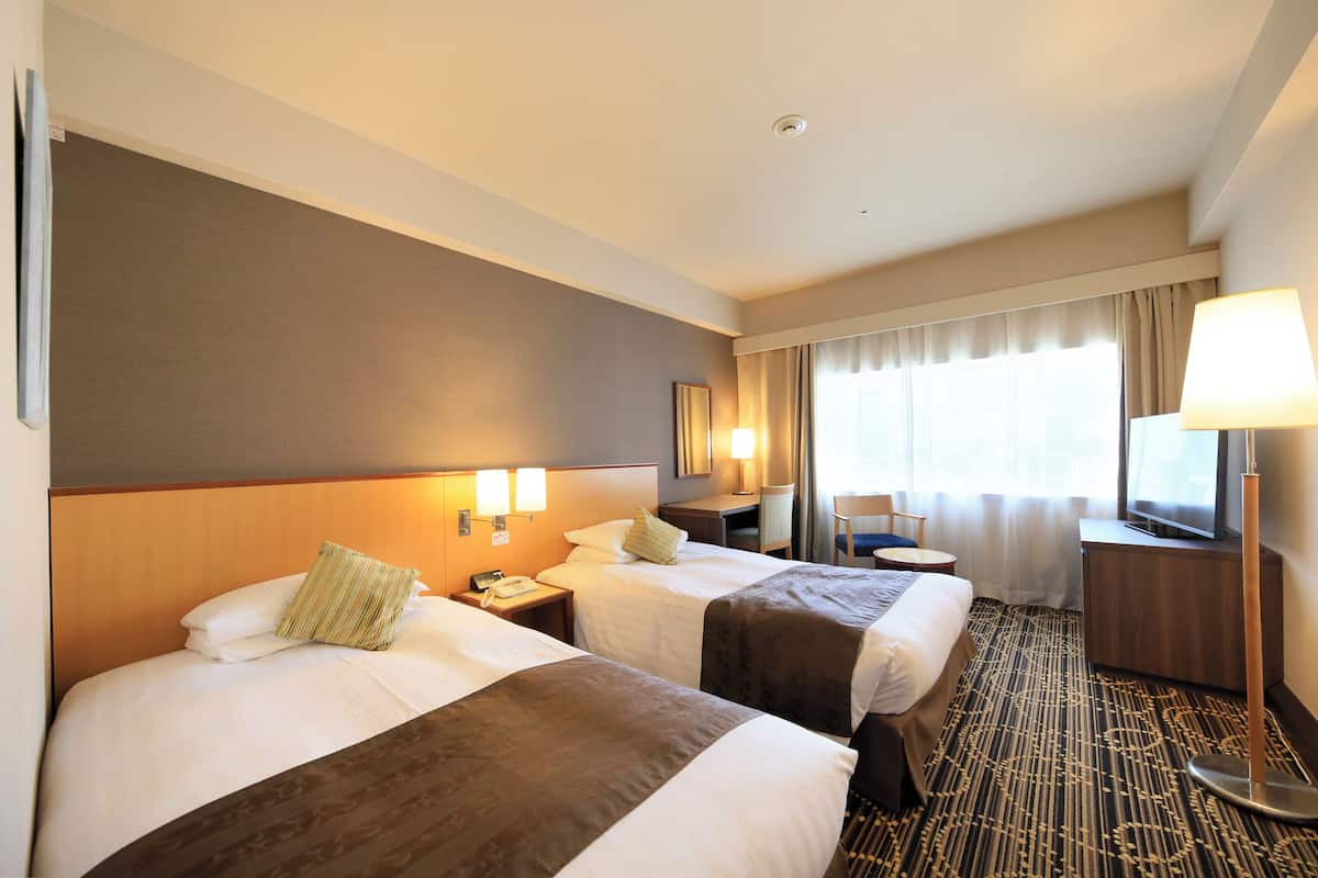Quarto de hotel com duas camas, uma escrivaninha, uma cadeira, uma televisão e uma janela com cortinas.