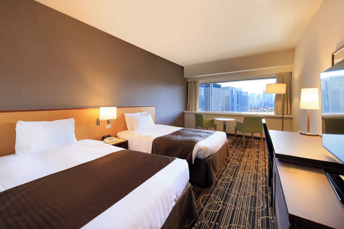 Quarto de hotel com duas camas, uma mesinha, uma cadeira e vista para a cidade.