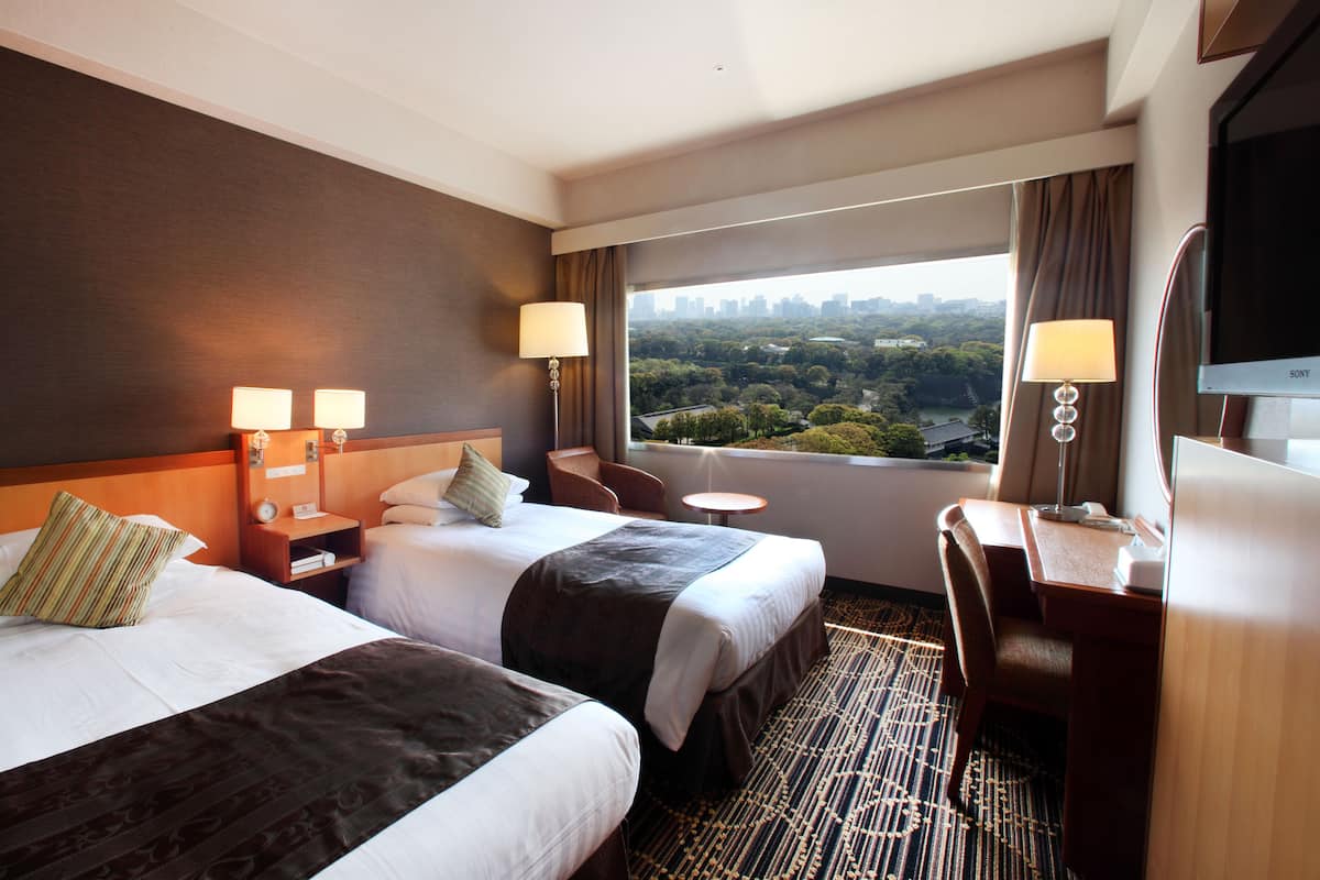 Quarto de hotel com duas camas, uma mesa, uma cadeira, uma televisão e vista para a paisagem urbana.