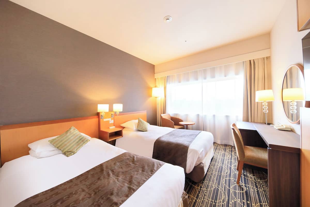 Quarto de hotel com duas camas, uma escrivaninha, uma cadeira, uma mesinha e um espelho.