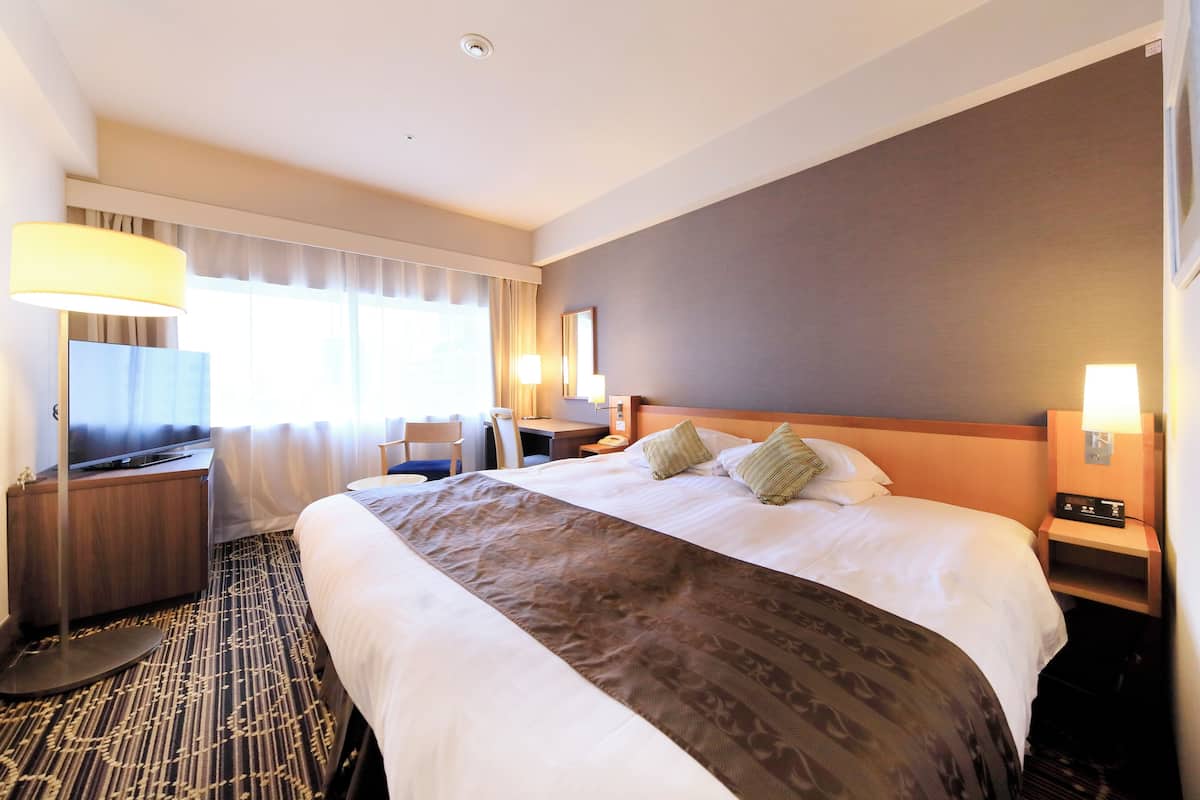 Quarto de hotel com uma cama grande, uma escrivaninha, uma cadeira, uma luminária de mesa, uma mesinha de cabeceira com abajur e uma janela com cortinas.