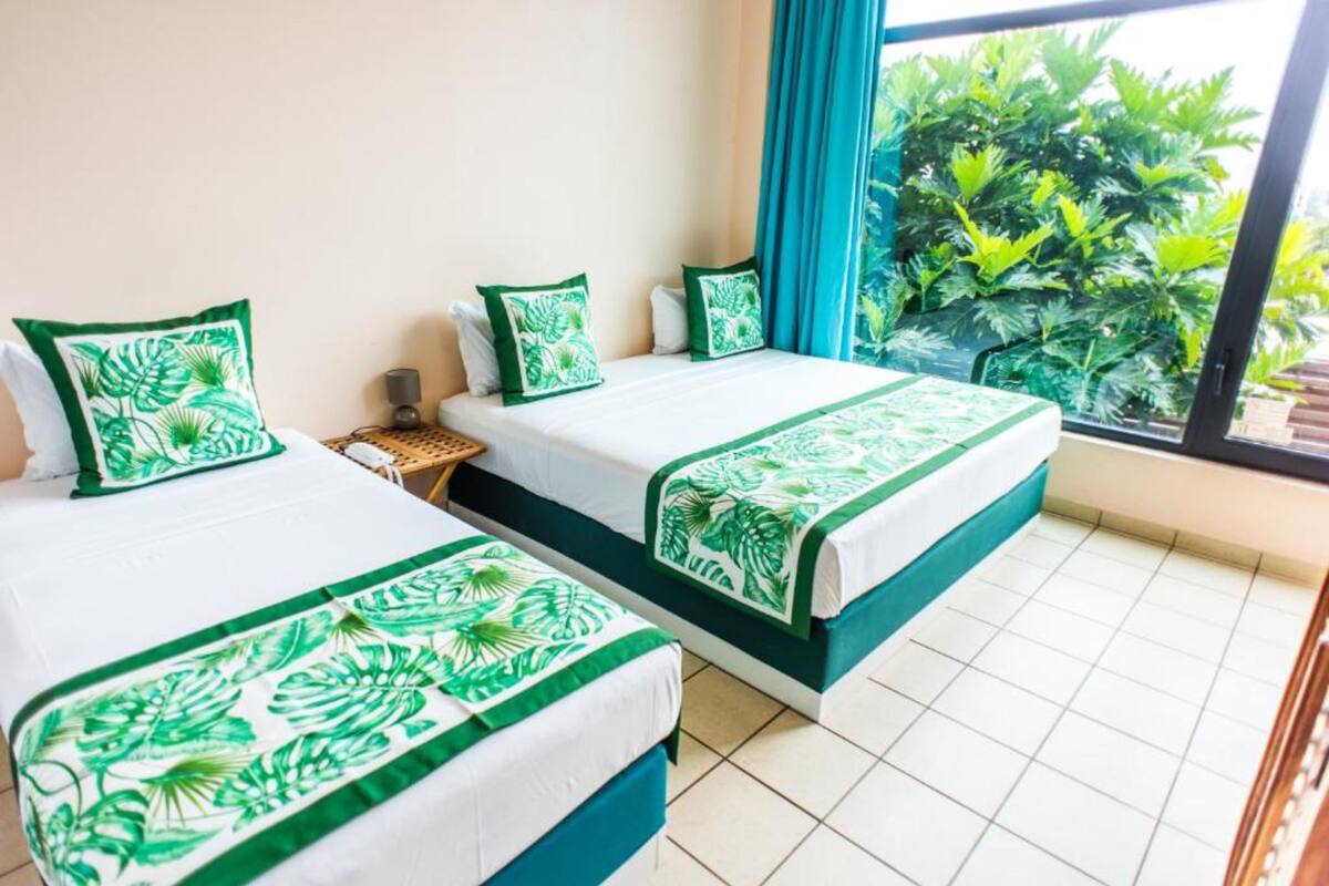 A hotel room with two beds, each with green leaf-patterned bedding, a small side table, and a view of lush greenery through a large window.