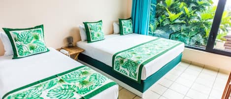 A hotel room with two beds, each with green leaf-patterned bedding, a small side table, and a view of lush greenery through a large window.