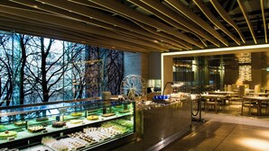 A modern hotel lobby with a dessert display, a large window with a view of trees, and a seating area with tables and chairs. - Nina Hotel Island South (Hong Kong)