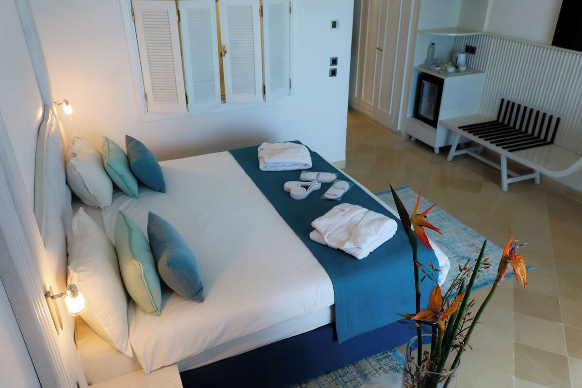 Una cama bien hecha con almohadas, una mesita de noche con una lámpara, un banco y un jarrón con flores.