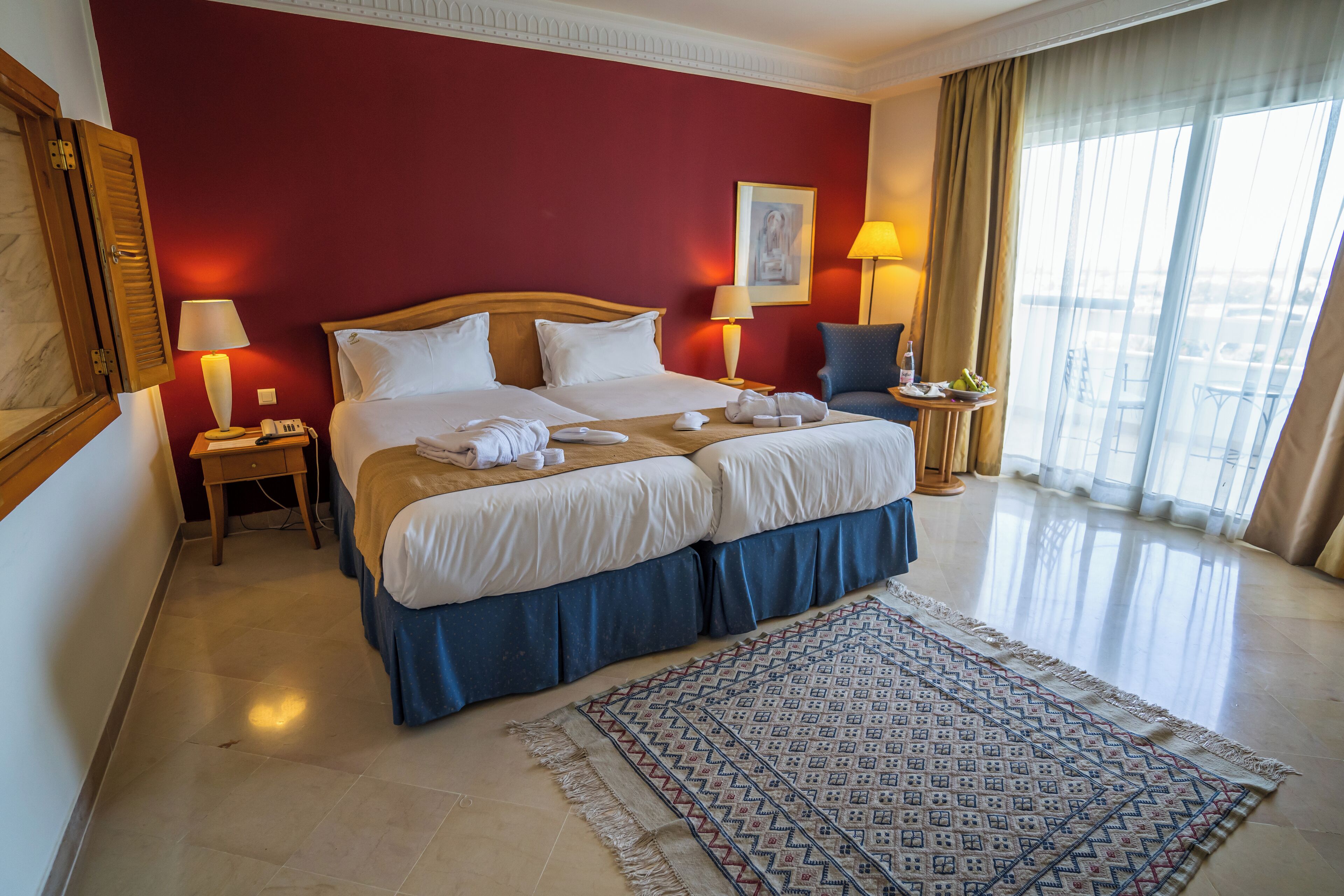 Een hotelkamer met een groot bed, twee nachtkastjes, een vloerkleed met dessin en uitzicht op de stad door grote ramen.