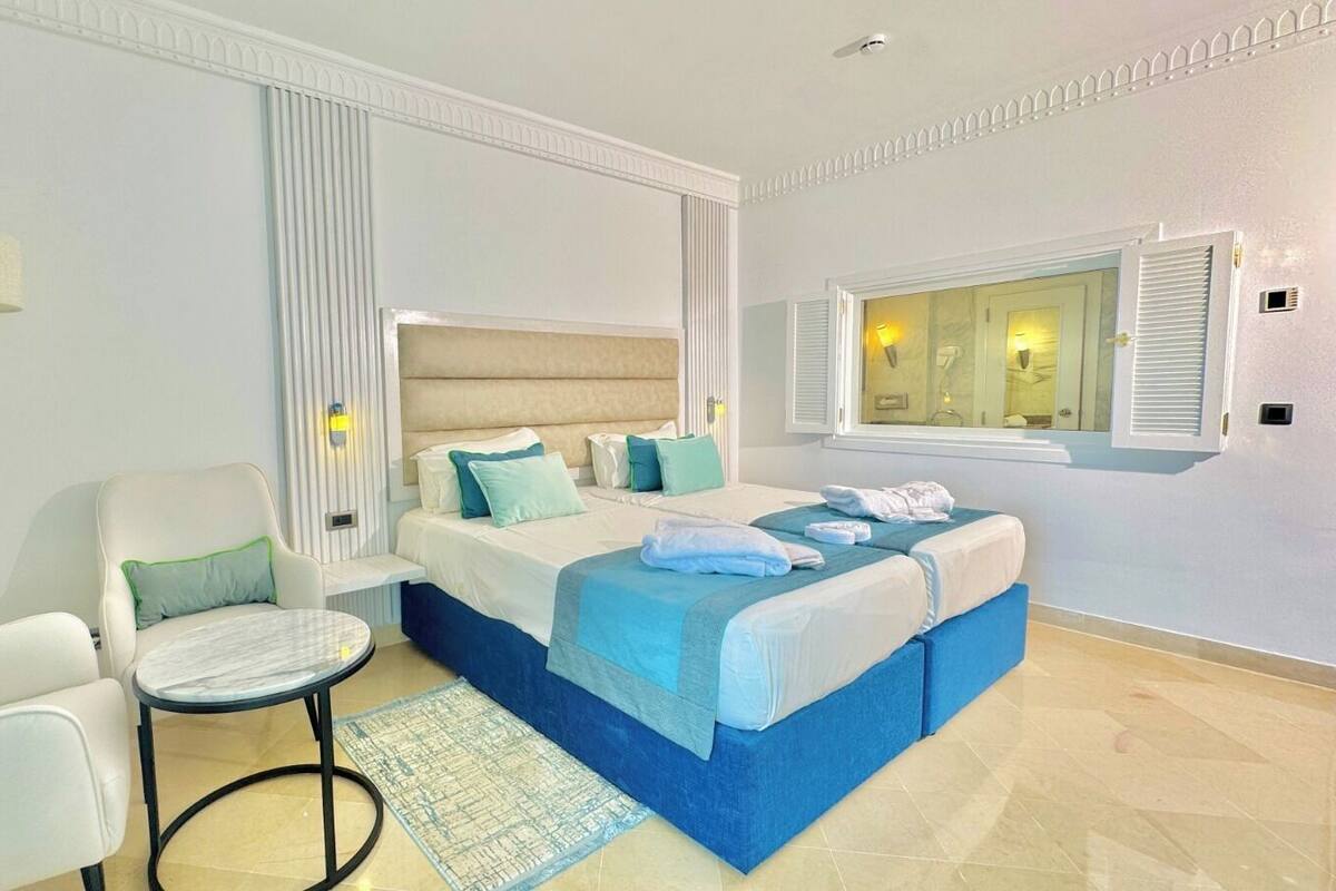 Habitación de hotel moderna con una cama grande, dos sillas, una mesita y un ventanal con persianas.