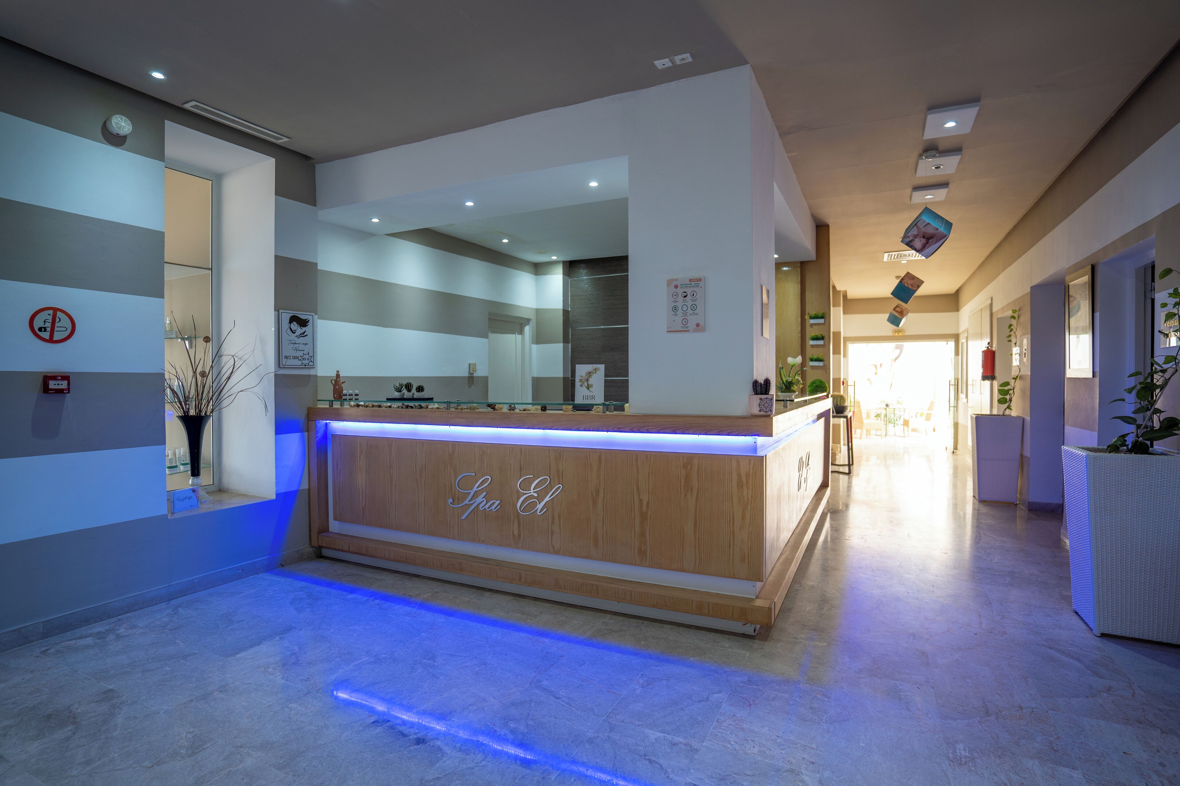 A reception area with a wooden counter labeled "Spa El," a blue-lit floor, and a hallway leading to a bright room.