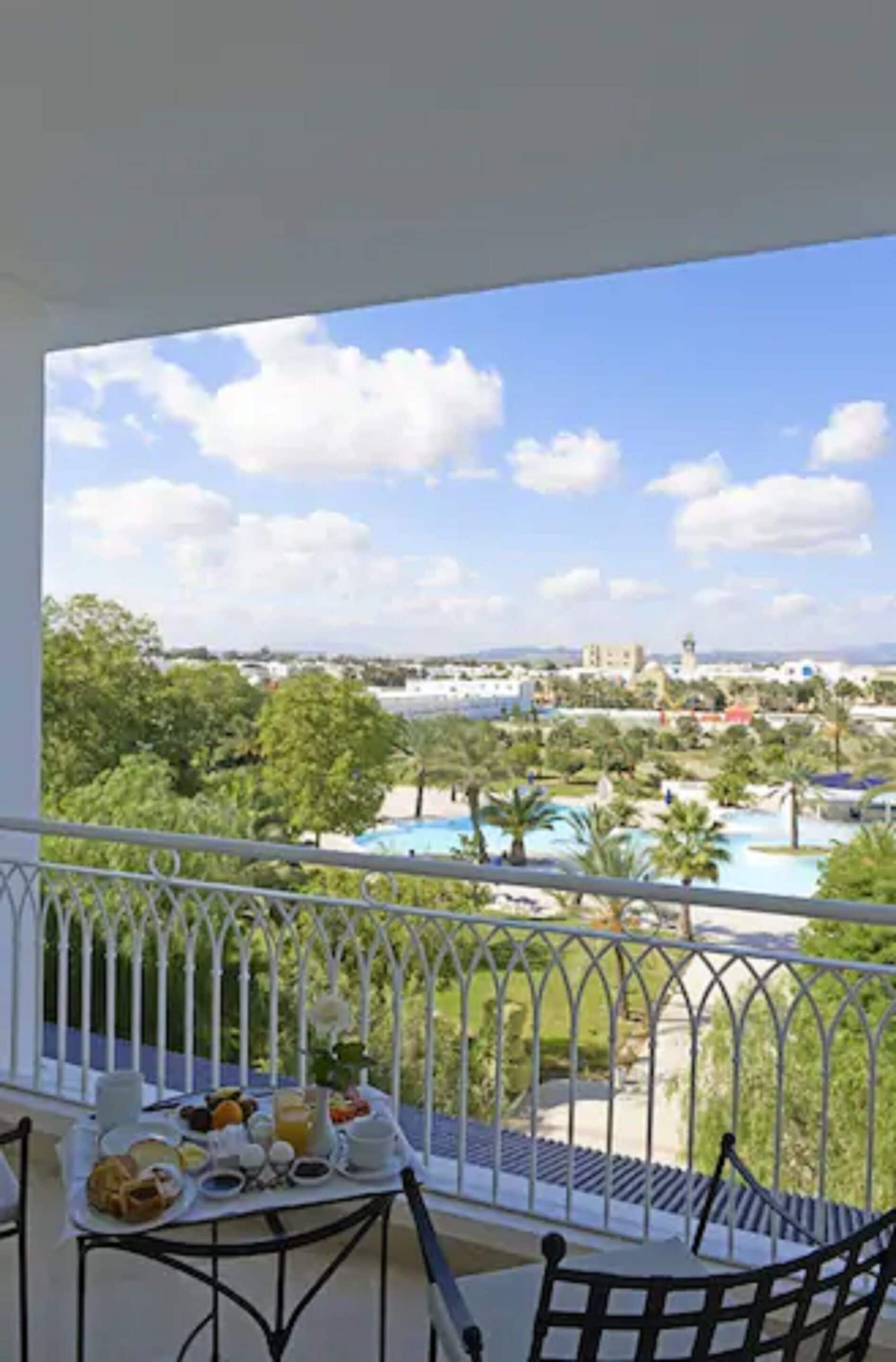 A balcony with a table set for breakfast, overlooking a pool and resort area.