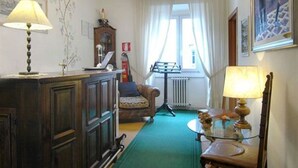 Um quarto com um armário de madeira, um tapete verde, uma cadeira, uma mesa com abajur, um radiador e uma janela com cortinas.