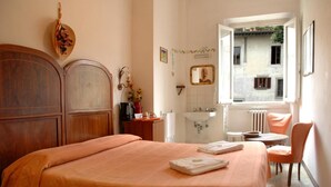 Quarto com cama de madeira, uma mesinha com cadeira, janela com vista para um edifício e banheiro com pia e espelho.