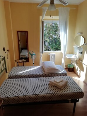 A room with two beds, a window with curtains, a radiator, a small table with a fan, a mirror, and a sink.
