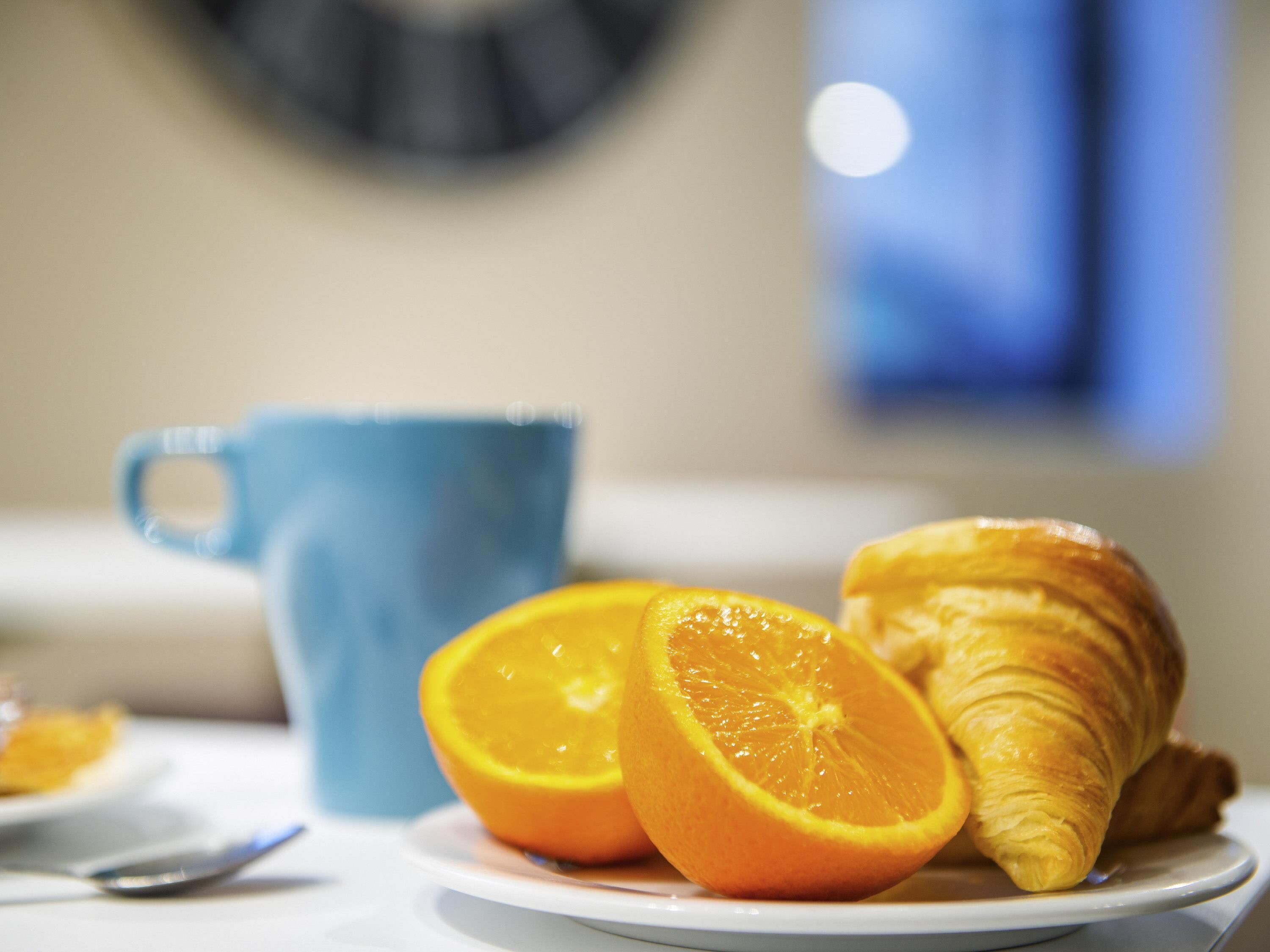 A plate with a croissant and an orange slice, a blue mug, and a spoon on a table.