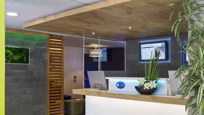 A reception area with a wooden reception desk, a computer, and a plant. There are white chairs and a grey wall with a green plant on the left. - ibis budget Cannes centre ville (Cannes)