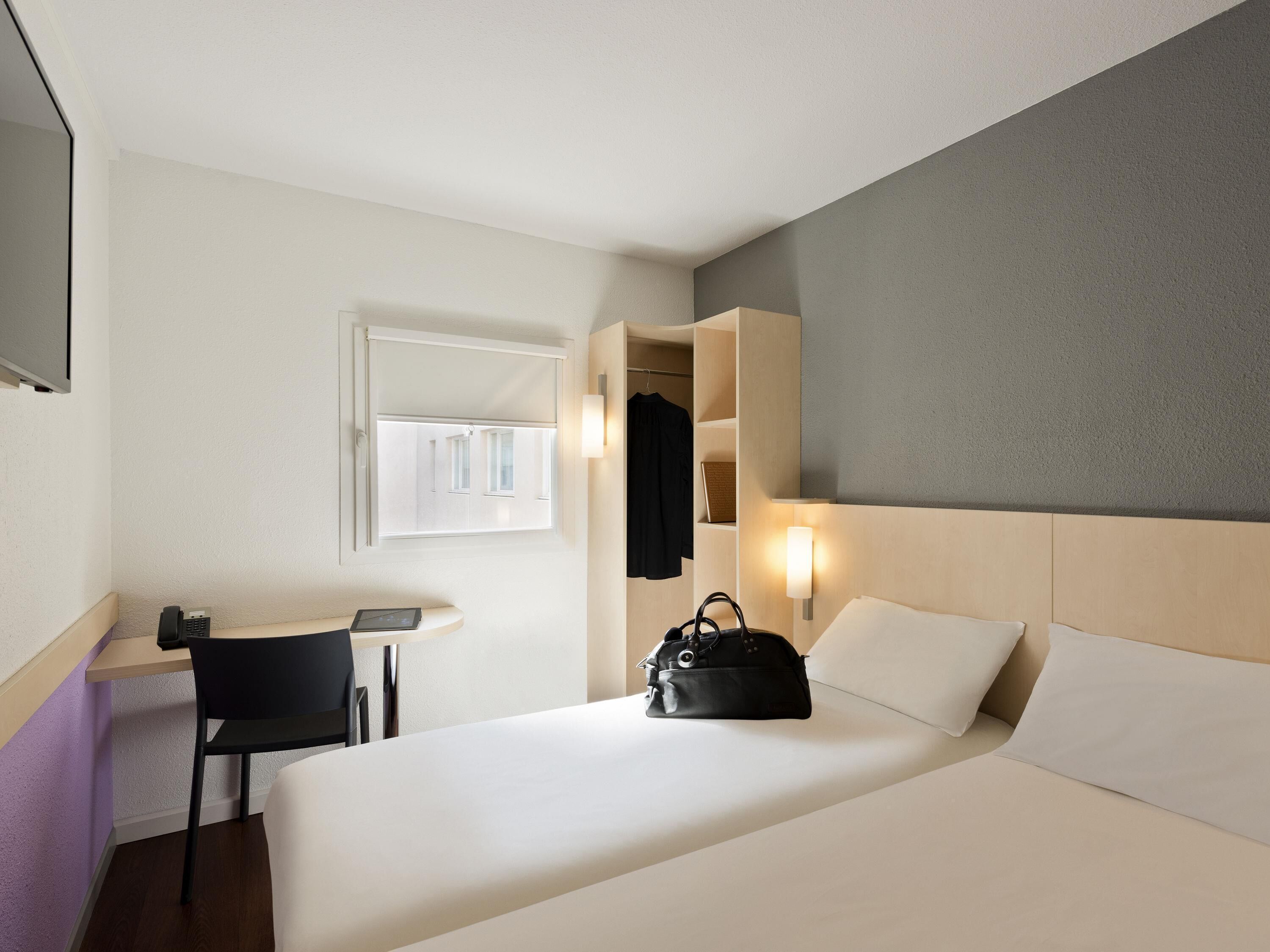 Une chambre d’hôtel avec un lit, un bureau, une chaise, un manteau accroché et une lampe fixée au mur.