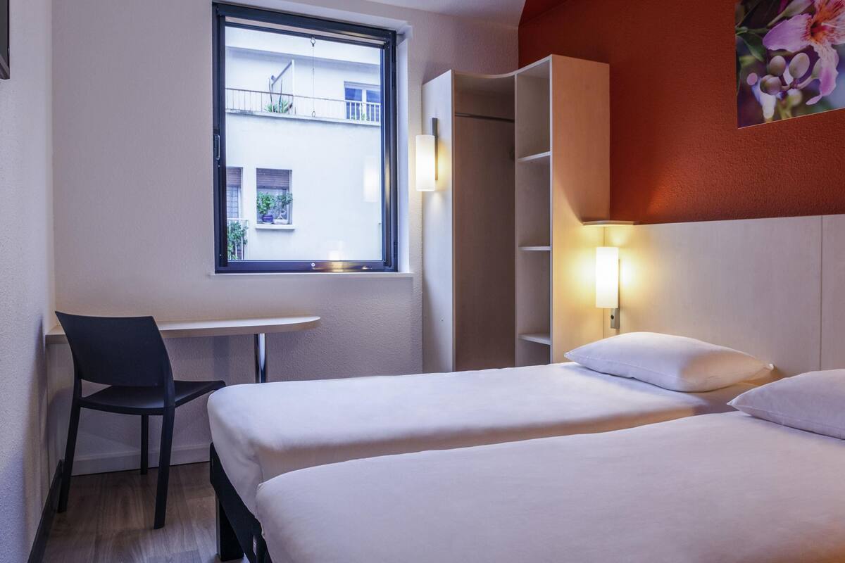 Hotelkamer met twee bedden, een bureau, een stoel, een raam met uitzicht op gebouwen en een wandlamp.