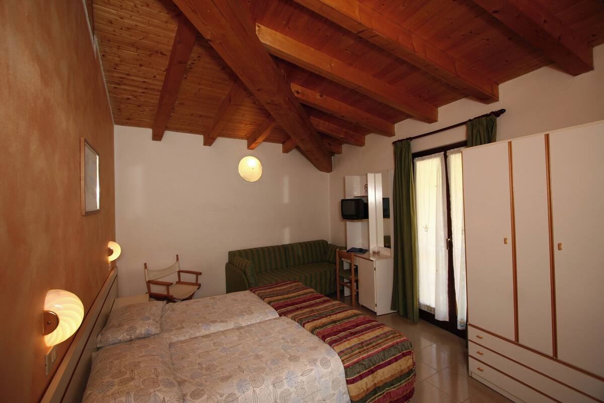Ein Schlafzimmer mit Holzbalkendecke, einem Bett mit gemusterten Betttextilien, einem grünen Sofa, einem Schreibtisch mit Stuhl, einem Kleiderschrank und einem Fernseher.