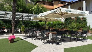 An outdoor patio with tables and chairs, covered by umbrellas, surrounded by greenery and buildings. - Stella D'Oro (Tremosine)