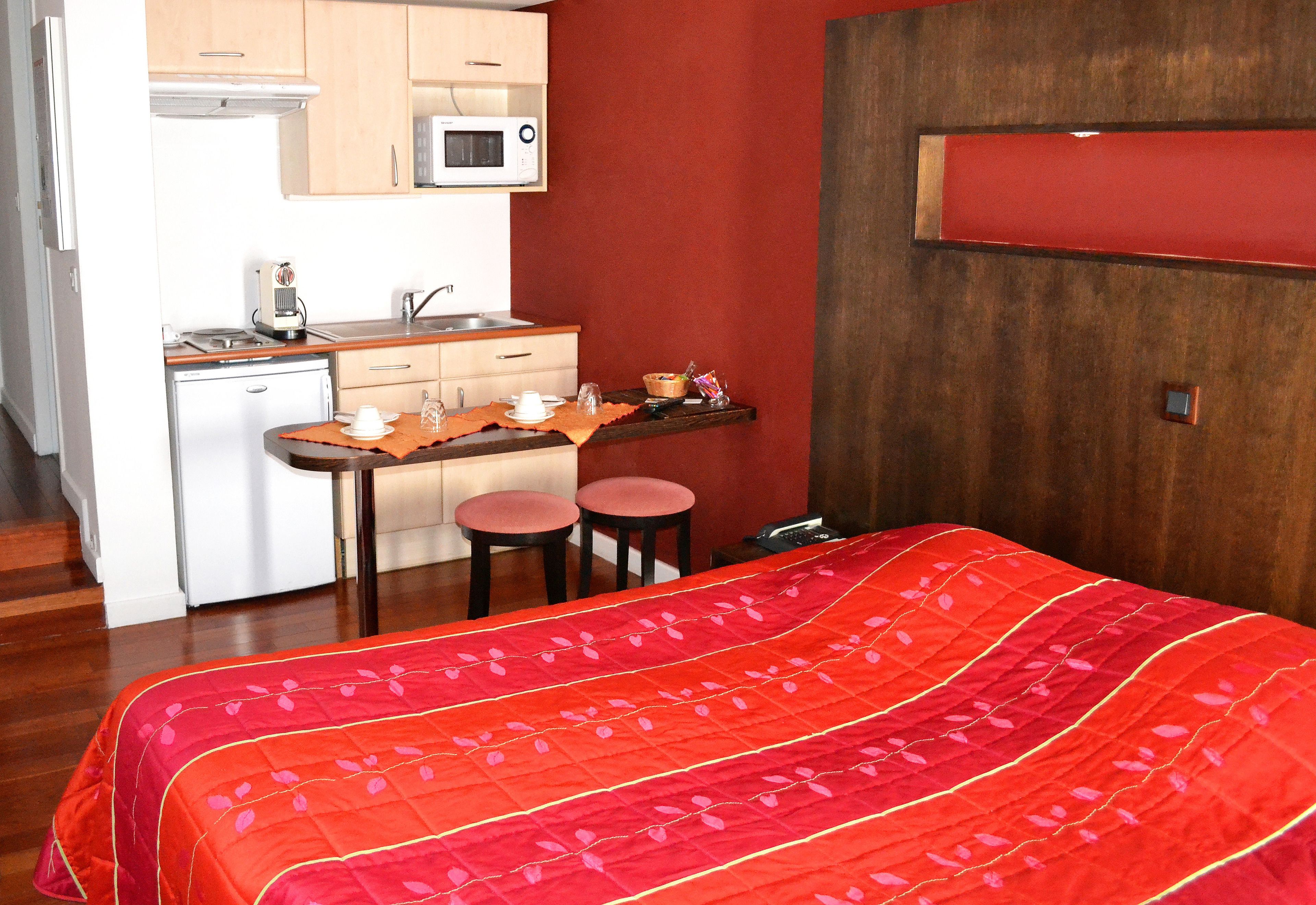 A compact hotel room with a kitchenette, a bed with a red quilt, and a wooden floor.