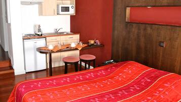 A compact hotel room with a kitchenette, a bed with a red quilt, and a wooden floor.