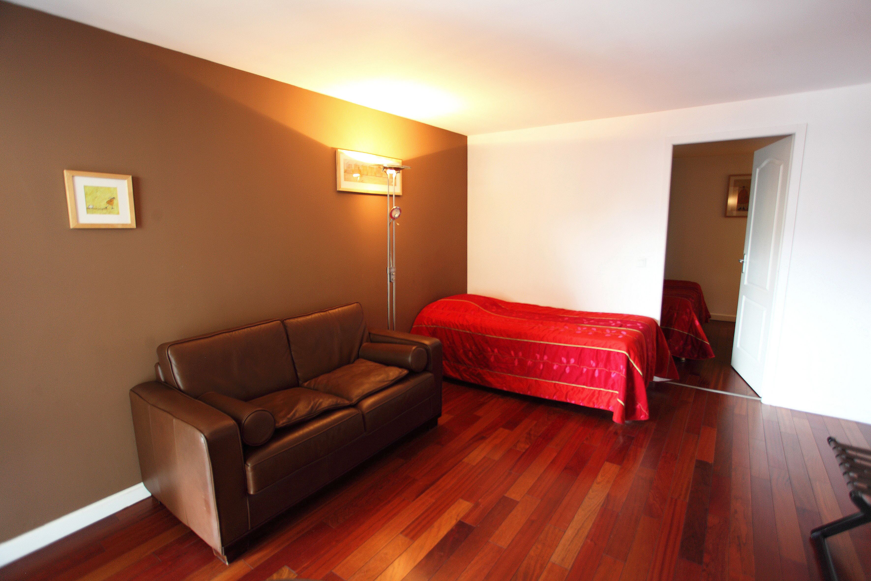 A room with a brown leather sofa, a red bed, and a floor lamp.