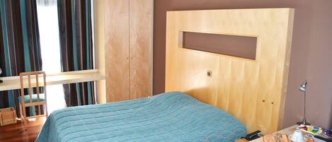 A bedroom with a bed, a wooden wardrobe, a desk with a chair, and a window with curtains.