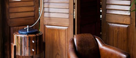 A wooden interior with a leather armchair, a round wooden side table with a lamp, and two framed pictures on the wall.