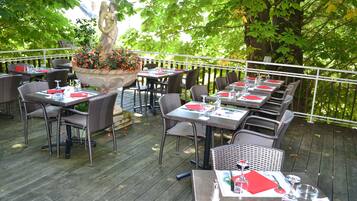 An outdoor dining area with wicker tables and chairs, a central planter with flowers, and a white railing.