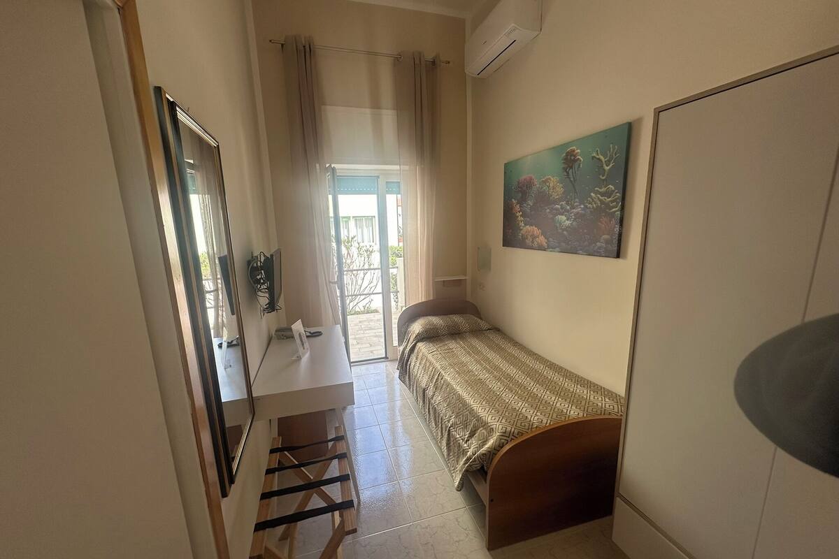 A small, well-lit room with a bed, a desk, and a chair. There is a window with curtains, a painting on the wall, and an air conditioning unit.