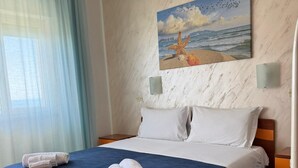 A hotel room with a bed, a painting of a beach scene, and a view of the sea through the window. - Hotel Germania (Praia a Mare)