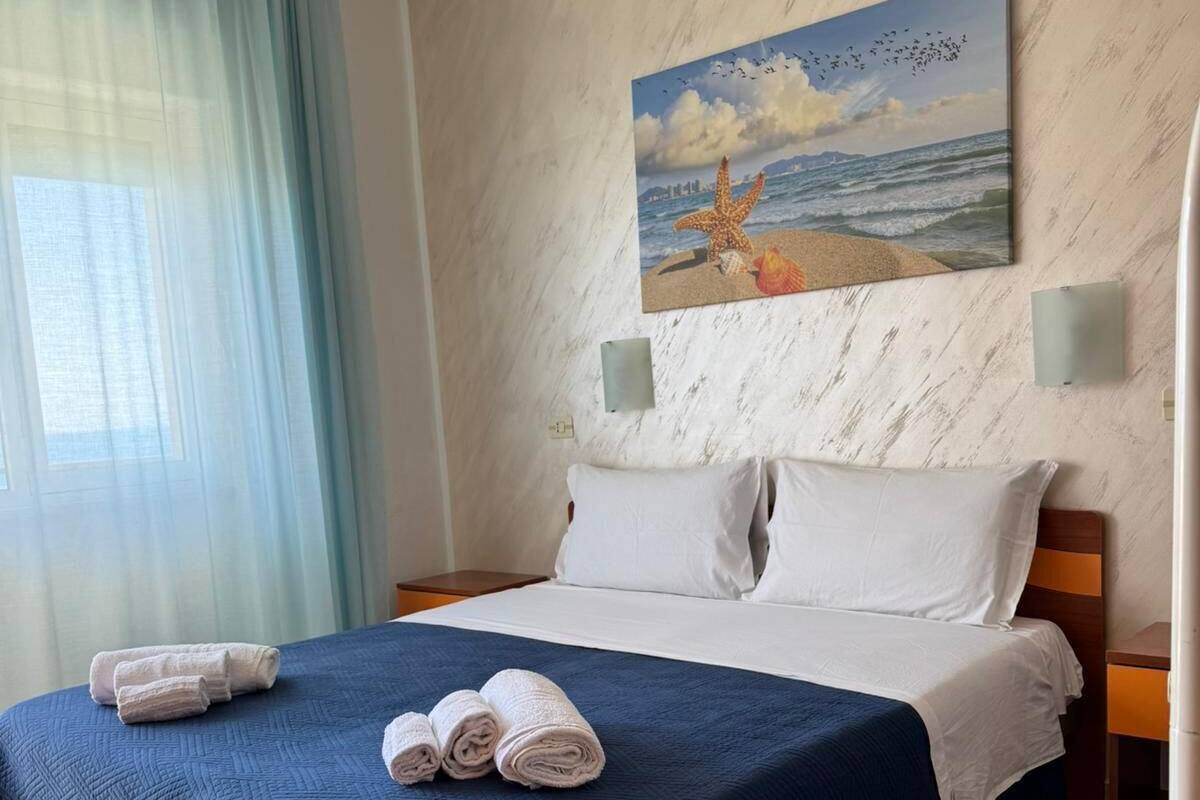 A hotel room with a bed, a painting of a beach scene, and a view of the sea through the window.