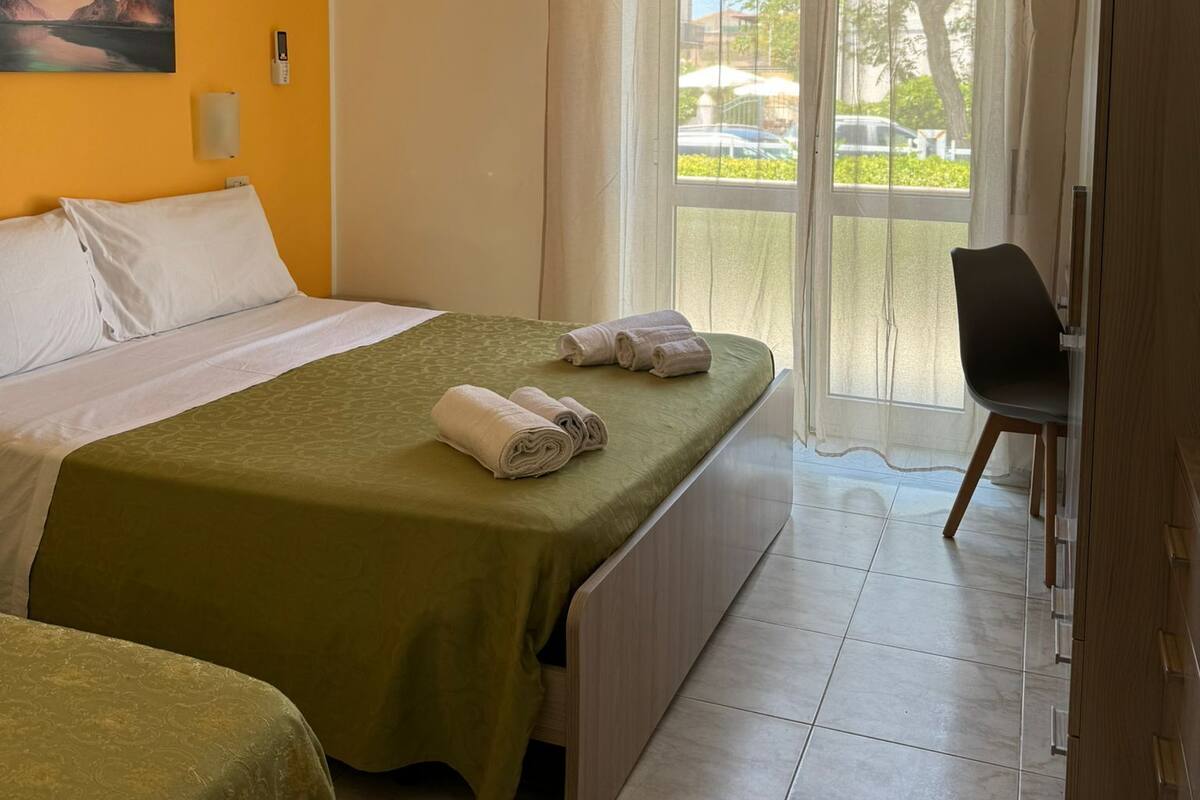 A hotel room with two beds, a painting on the wall, an air conditioner, a window with sheer curtains, and a chair by the window.