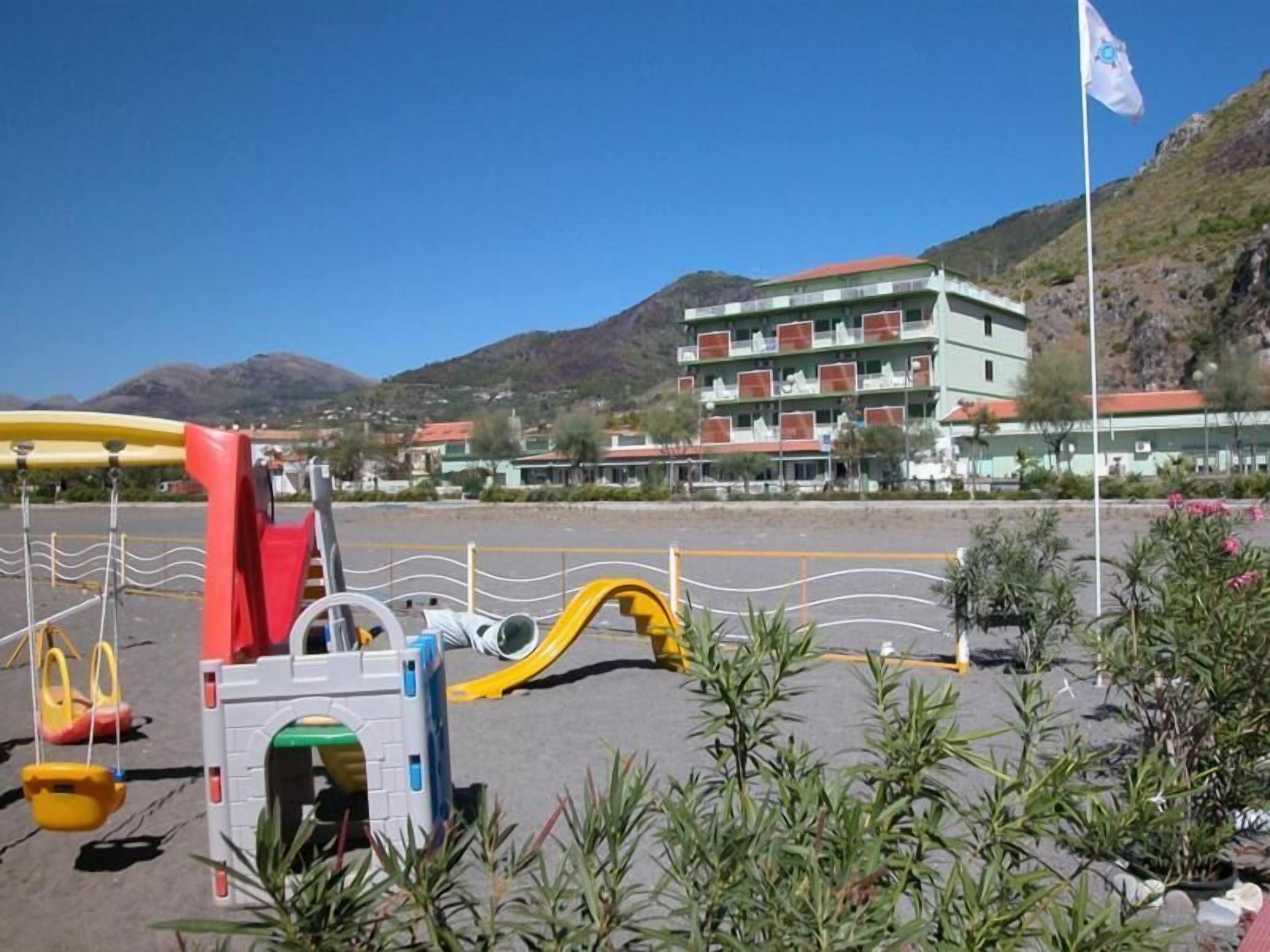 A playground with swings and slides, a building with balconies, and a flag in the background.