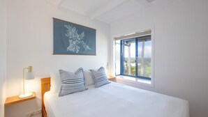 A bedroom with a large bed, a nightstand with a lamp, a window with a view, and a wall-mounted artwork.