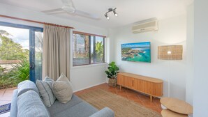 A modern living room with a sofa, a console table, a large window with blinds, a ceiling fan, an air conditioning unit, and a wall-mounted TV.