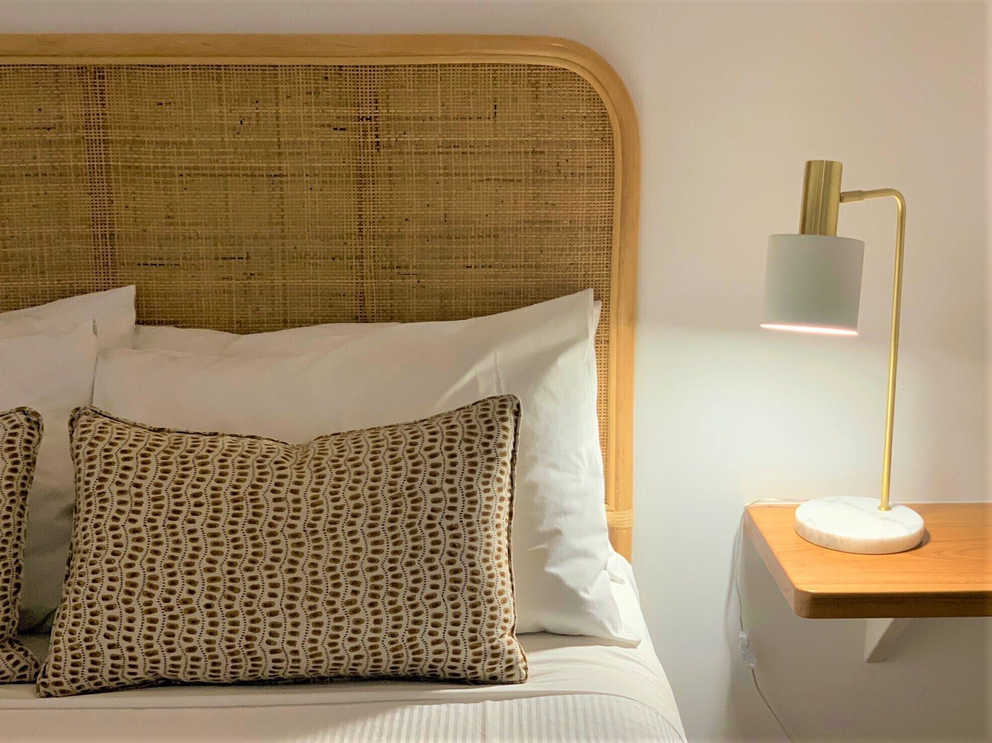 Ein Bett mit einem gewobenen Kopfteil, weißen Kissen und einem gemusterten Kissen. Außerdem ist ein Nachttisch mit einer Lampe zu sehen.