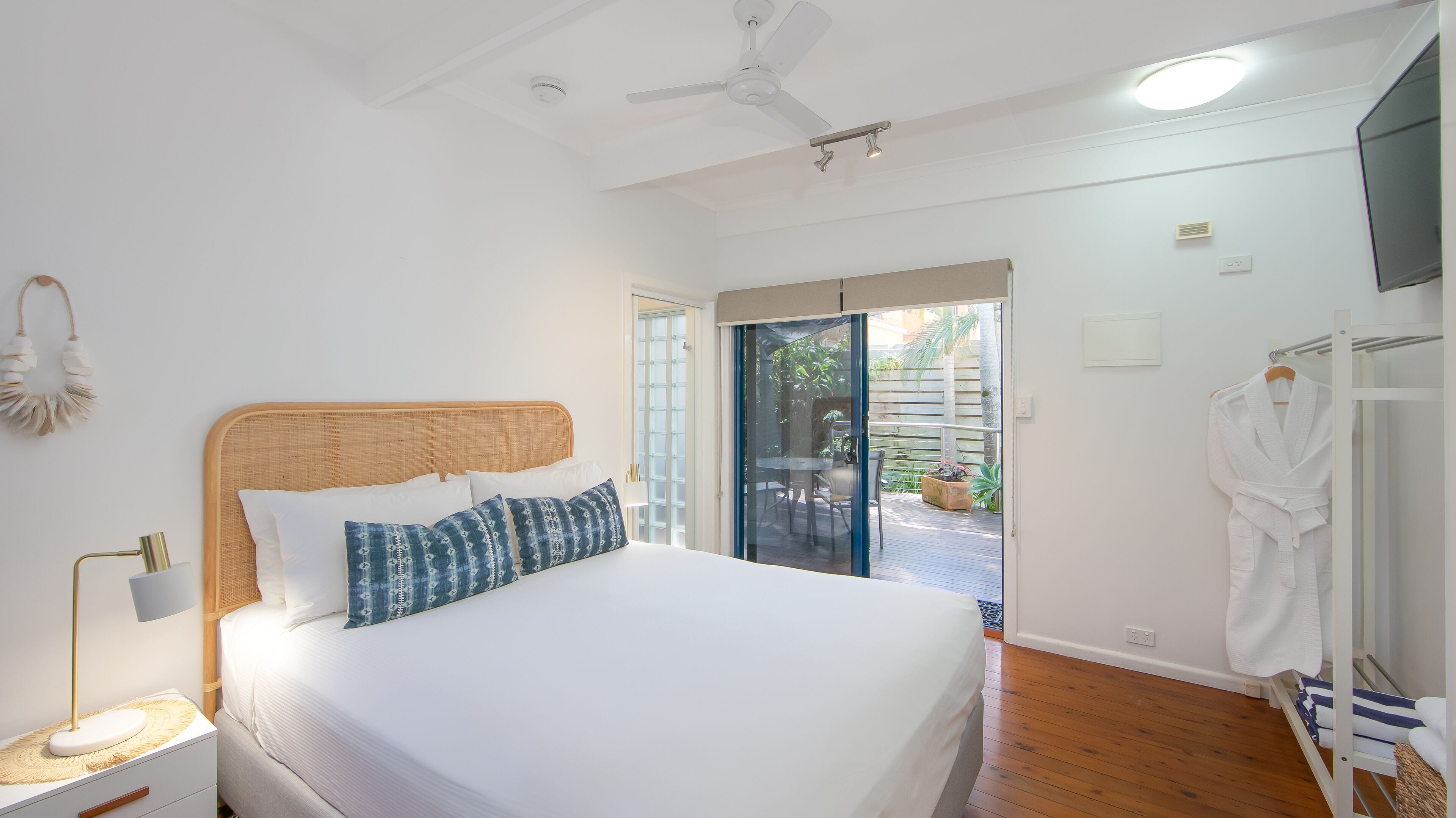 Ein Schlafzimmer mit Bett, Nachttisch, Lampe, Deckenventilator, einer Glastür zu einem Balkon und einem Bademantelständer.