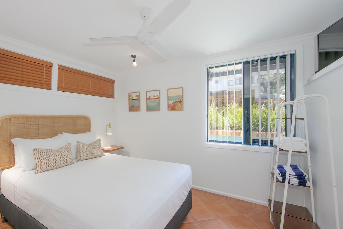 Ein Schlafzimmer mit einem Bett, Nachttischlampen, einem Deckenventilator, einem Fenster mit Jalousien und einer Leiter mit gefalteten Handtüchern.